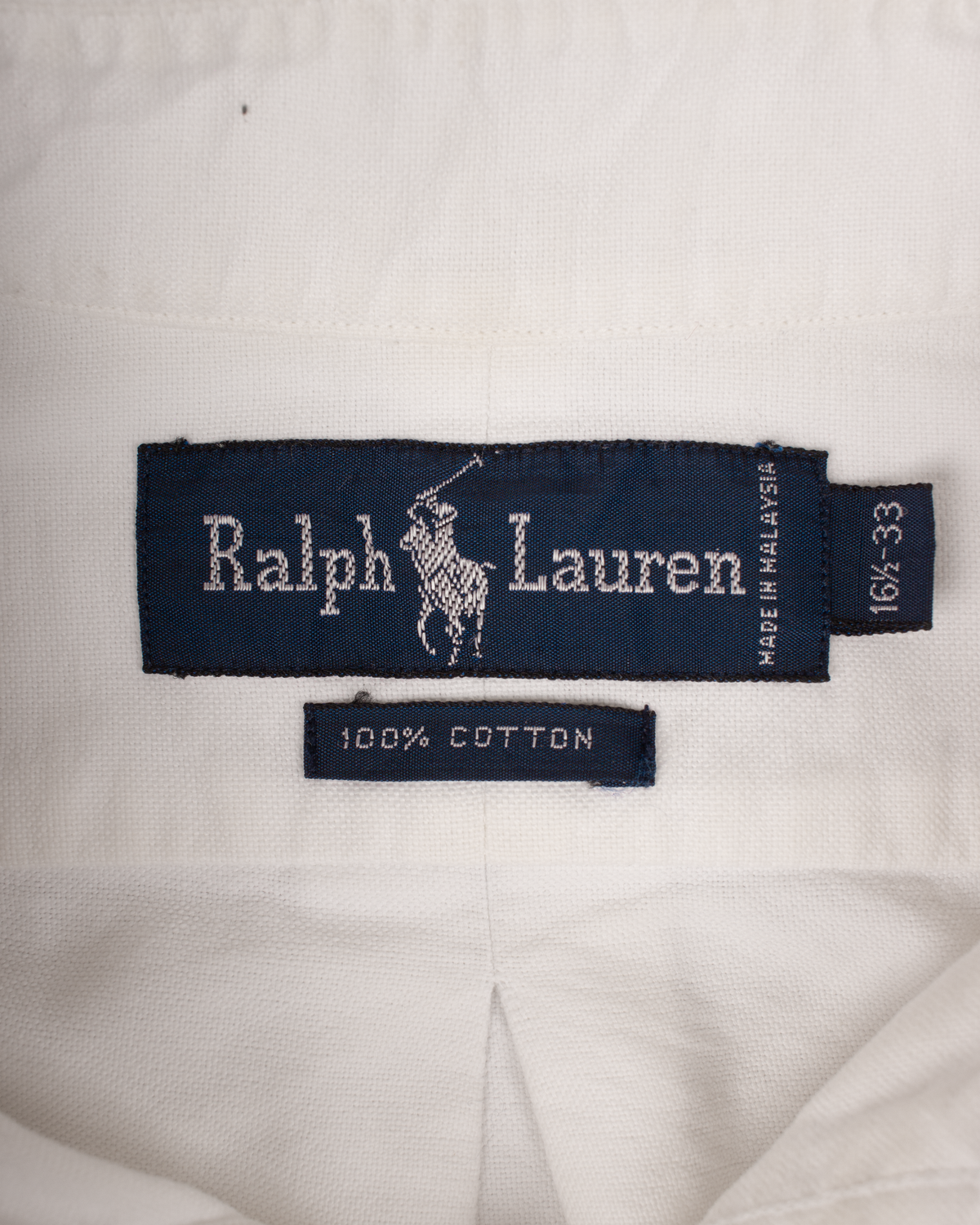 Close-up of a white Ralph Lauren garment showing the navy blue brand label with the white polo player logo and text, plus a smaller '100% COTTON' tag below.