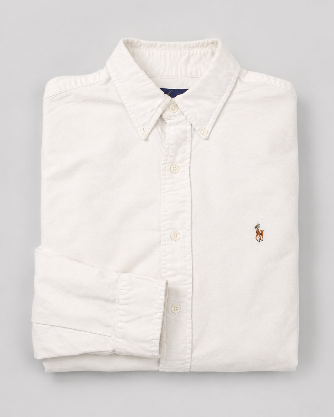 A folded white long-sleeve button-down shirt with a small embroidered polo player logo on the left chest, set against a neutral gray background.