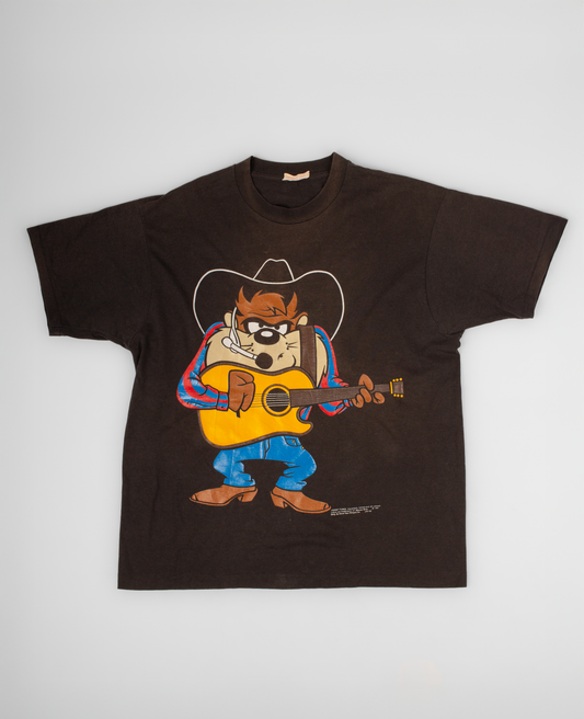 Brown t-shirt with cartoon character playing guitar on a white background Vintage Taz Tasmanian Devil tee featuring the beloved Looney Tunes character dressed as a country musician. During the 1990s, character graphic tees became a staple of American pop culture, with Taz standing out as one of the era’s most recognizable and collectible cartoon icons. A fun piece that blends vintage animation nostalgia with classic Americana style. single stitch

100% cotton, 1993, single stitched, made in USA, good fade